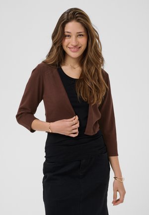 Femme aux longs cheveux bruns sourit, portant un cardigan marron sur un haut et une jupe noirs, debout devant un fond uni clair.