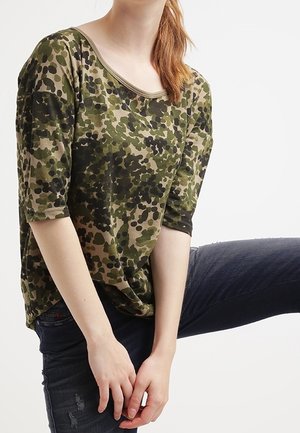 Haut en coton à imprimé camouflage en vert et beige, avec une coupe ample, un col large et des manches 3/4. Associé à un jean en denim foncé.