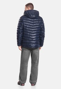 Granatowa kurtka puchowa o fakturowanym, błyszczącym wykończeniu, z elastycznym kapturem i przeszywanymi panelami. Połączona z luźnymi szarymi spodniami i ciemnym obuwiem.