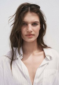 Portrait of a person with long, wavy brown hair, wearing an unbuttoned white striped shirt. Neutral background highlights facial features.