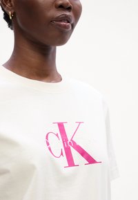 Kremkrāsas T-krekls ar īsām piedurknēm, kas rotāts ar lielu rozā "CK" logotipu un izšūtu "Calvin Klein" tekstu. Mīksts kokvilnas audums ar apaļu kakla izgriezumu.