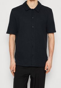 Schwarzes Kurzarmhemd mit Knopfleiste aus strukturiertem Stoff, mit Kragen und fünf Knöpfen vorne.