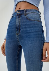 Kvinna som bär högmidjade blå denimjeans och en ljusblå kortärmad topp med volanger på ärmarna och nederkanten.