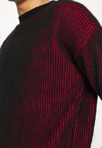 Pull rayé noir et rouge présentant un motif en tricot texturé, un col rond et des manches longues, avec des couleurs alternées distinctes.