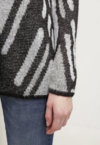 Sweter dziergany w czerni i szarości, z wyrazistymi, abstrakcyjnymi wzorami. Długie rękawy o teksturowanym wykończeniu i ściągacz na dole.