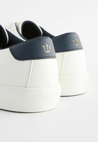 Weiße Sneakers mit strukturiertem Finish, ausgestattet mit einer marineblauen Fersenplatte und einem kleinen goldenen Krone-Logo. Gummisohlen für besseren Halt.