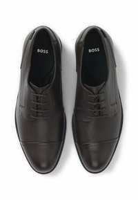 Scarpe in pelle marrone con finitura testurizzata, dotate di un design con lacci, punta arrotondata e dettagli di cucitura discreti. Logo del brand visibile all'interno.