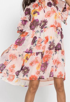 Femme portant une robe légère et fluide avec des imprimés floraux violets, orange et jaunes, tenant la robe déployée d'un côté.