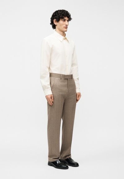 Άνδρας που στέκεται φορώντας κρεμ πουκάμισο με μακριά μανίκια και κουμπιά, μπεζ παντελόνι με ρίγες και ζώνη, και μαύρα επίσημα παπούτσια σε λευκό φόντο.