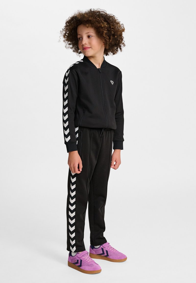 Zwarte trainingsoutfit met witte chevronstrepen langs de mouwen en benen, ritssluiting jacket en zwarte broek. Gecombineerd met paarse sneakers.