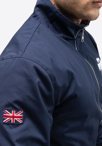 Marineblå jakke med høj krave, udstyret med en sølv lynlås og et broderet britisk flag-patch på venstre ærme. Glat, slidstærkt materiale.