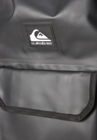 Svart vanntett bag med teksturert overflate, utstyrt med et innsydd logo-patch. Inkluderer en frontlomme med svart kant og robust maskinvare.