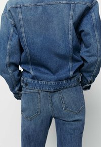 Farkkutakki ja farkut keskisinisessä pesussa. Ominaisuuksiin kuuluu klassinen muotoilu nappikoristeilla, etupockett ja näkyvät tikkaus yksityiskohdat.