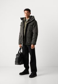 Giacca isolante verde scuro con molteplici tasche, chiusura con zip e cappuccio removibile, abbinata a un maglione verde chiaro con zip e pantaloni neri.
