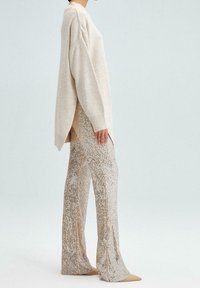 Beige oversized pletený svetr s bočními rozparky, spárovaný s lesklými flitrovými kalhotami v zlatých tónech a nazouvacími béžovými lodičkami s špičatou špičkou.