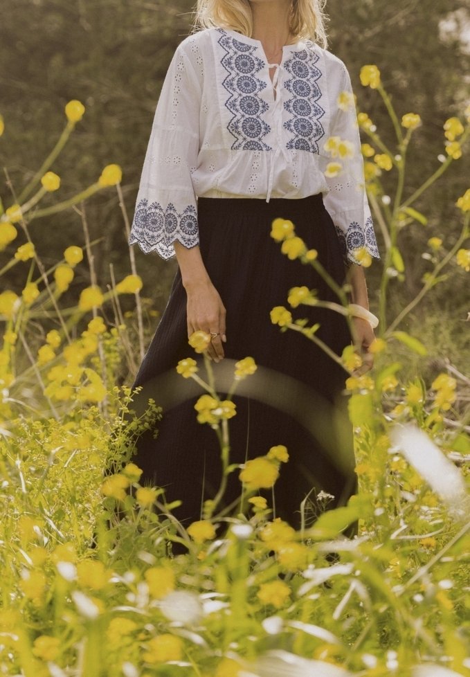 Personne en blouse blanche brodée et jupe noire debout dans un champ de hautes fleurs sauvages jaunes, en plein air avec des arbres verts en arrière-plan.