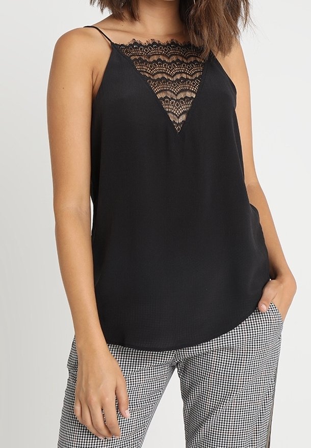 Femme portant un caraco noir avec un décolleté en V en dentelle et de fines bretelles, associé à un pantalon à carreaux noir et blanc.