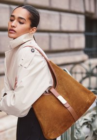 Brun ruskind tote taske med glatte læderdetaljer og en magnetisk lukning. Har en struktureret form og kontrasterende lysebrune håndtag.