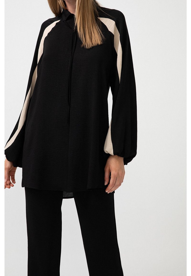 Femme portant une tunique noire ample avec de larges rayures crème le long des manches et un pantalon noir assorti sur un fond uni.