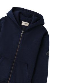 Sudadera con cremallera azul marino hecha de tela suave. Cuenta con una gran capucha, bolsillos frontales y un parche con un logo triangular en la manga.