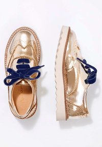 Scarpe metallizzate dorate con finitura liscia, caratterizzate da lacci blu, dettagli brogue e suola bianca seghettata.