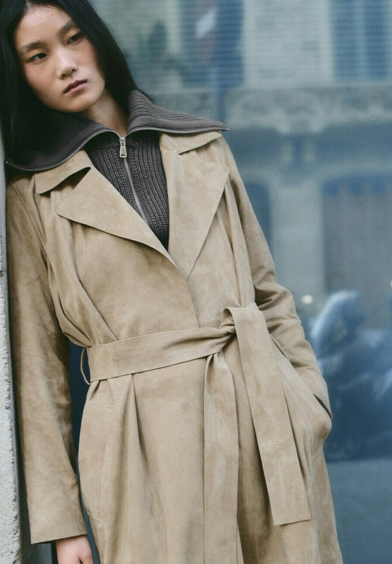Donna appoggiata a un muro che indossa un cappotto trench beige con cintura sopra un maglione marrone con cerniera, con capelli lunghi e neri e un'espressione neutra.