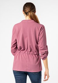 Haut rose à manches longues avec un effet portefeuille, taille froncée et manches retroussables. Matière douce avec une texture lisse et un design simple et épuré.