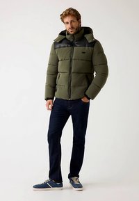 Giubbotto imbottito verde oliva con pannelli neri sulle spalle, chiusura frontale con zip, trama imbottita e cappuccio. Abbinato a jeans scuri e sneakers blu.
