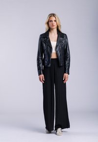 Ung kvinna står, klädd i svart läderjacka, vit kort topp, vida svarta byxor och vita sneakers mot en enfärgad grå bakgrund.