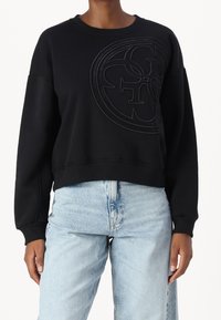 Sudadera negra con un logo texturizado y tonal en el centro; cuenta con cuello redondo y mangas largas; combinada con jeans de mezclilla azul claro.