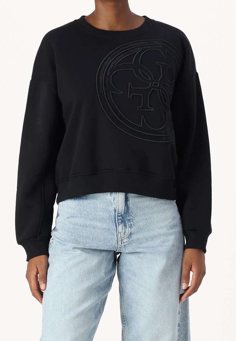 Sudadera negra con un logo texturizado y tonal en el centro; cuenta con cuello redondo y mangas largas; combinada con jeans de mezclilla azul claro.