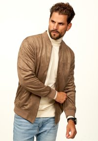 Braune Wildleder-Bomberjacke mit Reißverschluss vorne, gerippten Bündchen und strukturiertem Finish, getragen über einem beigen Rollkragenpullover, kombiniert mit hellen Jeans.