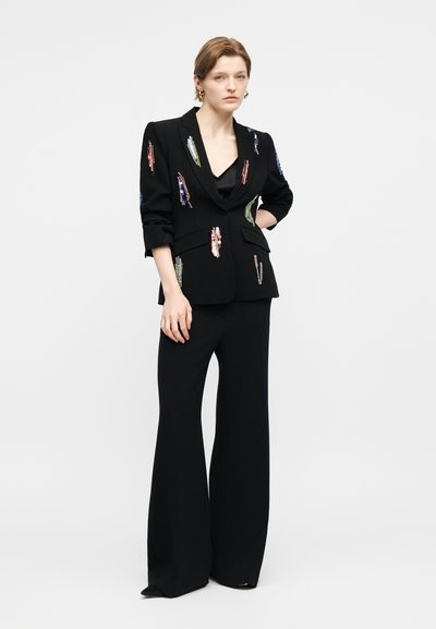 Γυναίκα που φοράει μαύρο ραμμένο σακάκι με πολύχρωμα παγιετένια μπαλώματα, μαύρο παντελόνι με φαρδιά γραμμή και μικρά στεφάνια σκουλαρίκια, στέκεται με το χέρι στη μέση.