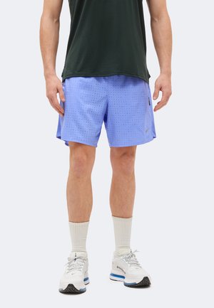 Man draagt lichtpaarse geperforeerde sportshorts, witte sokken, witte hardloopschoenen en een zwart mouwloos topje, staand tegen een witte achtergrond.