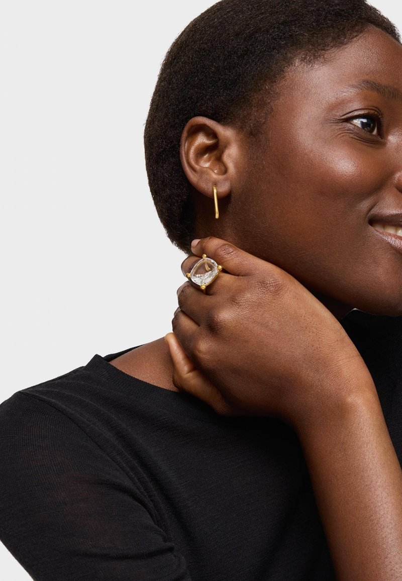 Złota kolczyk koło i duży pierścionek z przezroczystym kamieniem. Modelka ma krótkie włosy, ubrana w czarny top na jasnym tle.