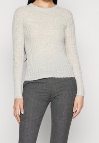 Donna che indossa un maglione a coste beige a maniche lunghe e pantaloni grigi a motivo herringbone, in piedi di fronte a uno sfondo semplice.