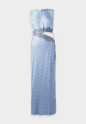 Femme debout dans une robe maxi bleue sans manches à motifs, avec une découpe et un détail décoratif à la taille, portant des talons hauts noirs.