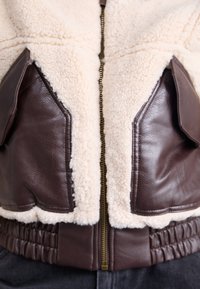 Giacca in pelle marrone e sherpa crema con chiusura a zip, cintura a coste e tasche a patta con accenti lisci.