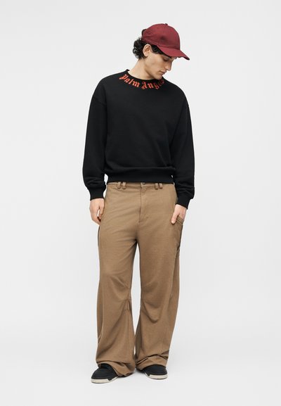 Νέος άνδρας με μαύρο φούτερ με κόκκινα γράμματα, μπεζ παντελόνι με φαρδιά μπατζάκια, μαύρα αθλητικά παπούτσια και κόκκινο καπέλο του μπέιζμπολ, που κοιτάζει προς τα κάτω.