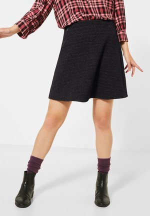 Woman wearing black textured A-line skirt, pink and black plaid long-sleeve shirt, purple socks, and black ankle boots standing with legs apart.