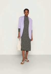 Blazer lilás claro sobre um vestido preto com padrão floral branco, acompanhado de uma mala creme e sandálias brancas com tiras de tornozelo.
