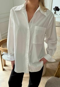 Fehér, galléros ing hosszú ujjakkal és mellzsebbel. Könnyű anyagból készült, enyhén gyűrött textúrával.