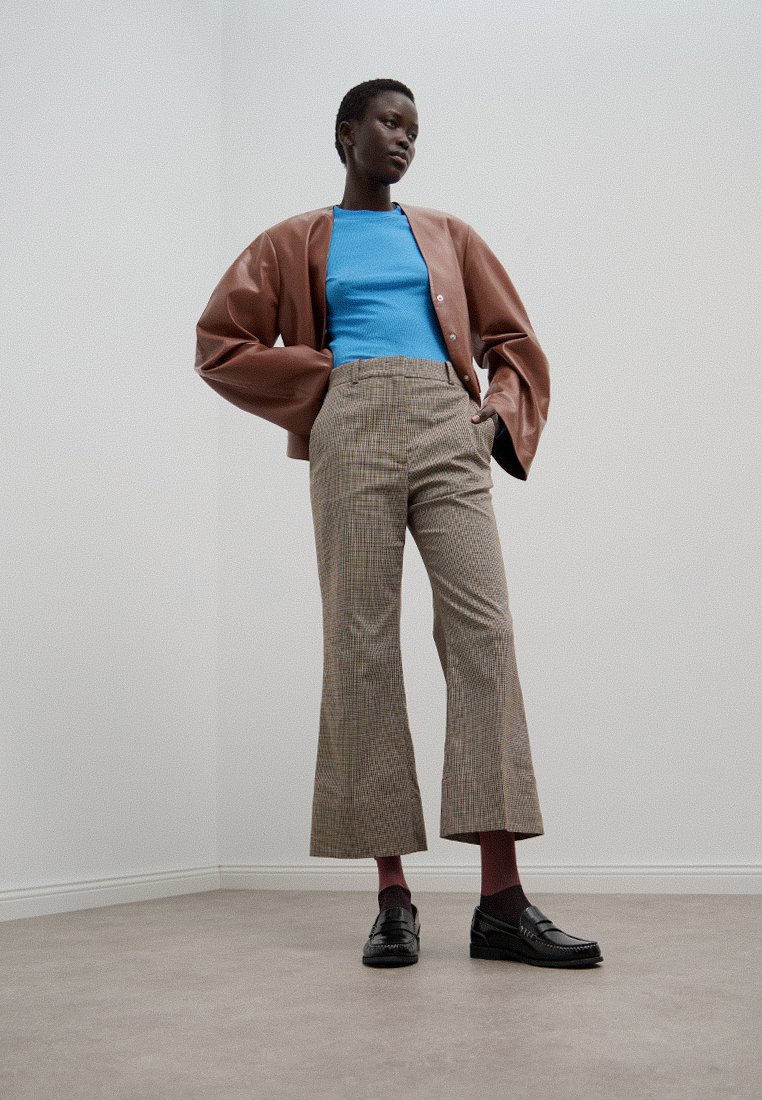 Brązowa kurtka z ekoskóry w oversize, niebieska dopasowana bluzka, wzorzyste spodnie w kratę z wysokim stanem, czarne mokasyny i bordowe skarpetki. Gładkie tło.