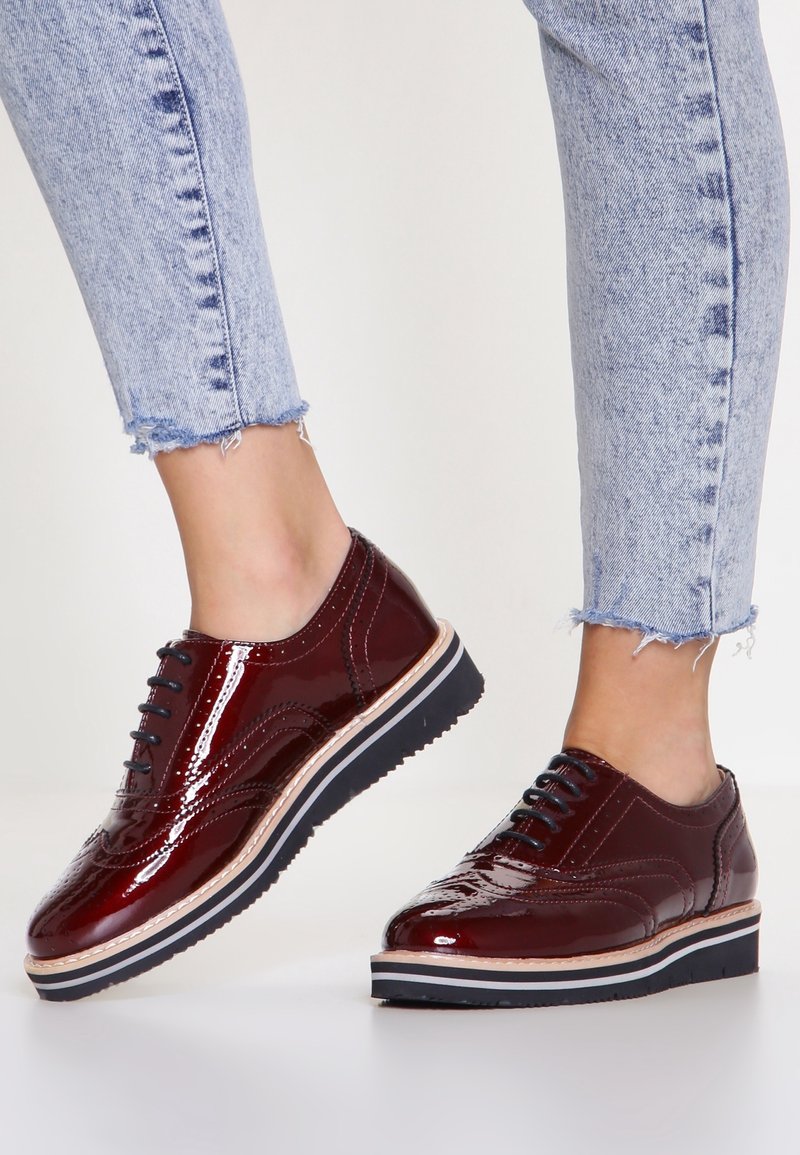 Personne portant des chaussures brogues bordeaux brillantes avec des lacets noirs et un jean bleu clair court debout sur un sol blanc.
