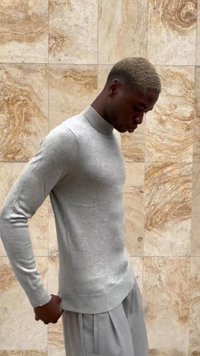 Maglione grigio chiaro con colletto a girocollo a coste, maniche lunghe e silhouette aderente, abbinato a pantaloni grigi, su uno sfondo strutturato.