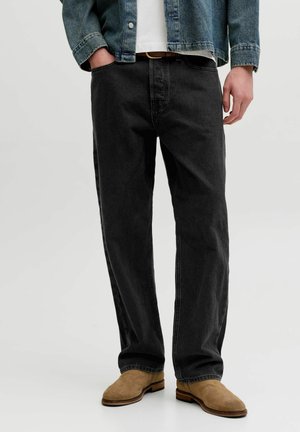 Muž nosiaci čierne rifle s rovnými nohavicami, béžové semišové topánky, bielu košeľu a modrú rifľovú bundu, s jednou rukou v vrecku.