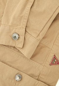 Manche et poignet d’une veste en jean beige montrant des boutons métalliques et un patch logo triangulaire rouge de la marque Guess.