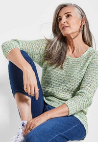 Strickpullover in Grün- und Gelbtönen mit U-Boot-Ausschnitt und 3/4-Ärmeln, kombiniert mit blauen Jeans und weißen Turnschuhen.