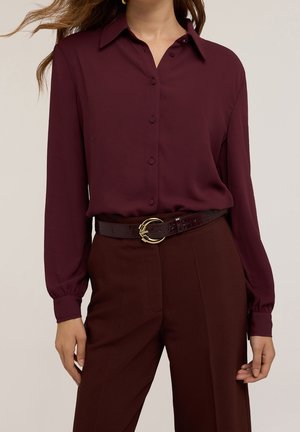 Kobieta nosząca bordową, długą bluzkę na guziki z długim rękawem, wpuszczoną w dopasowane, wysokie spodnie z ciemnym paskiem i złotą klamrą.