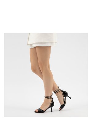 Beine, die weiße Shorts und schwarze High Heels mit offenem Zehenbereich und Knöchelriemen tragen, stehen vor einem einfarbigen weißen Hintergrund.